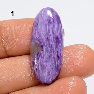 Puede incluir: Una gema pulida de forma ovalada con un patrón arremolinado de lavanda y blanco. La piedra tiene una superficie lisa y brillante y una pequeña inclusión gris claro. La gema se sostiene en una mano.