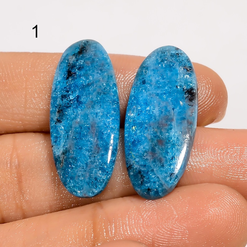 Puede incluir: Dos piedras preciosas ovaladas pulidas. Las piedras son de un vibrante azul turquesa con motas e inclusiones oscuras. Las piedras preciosas se muestran sobre un fondo neutro.