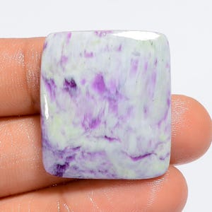Puede incluir: Un cabuj&oacute;n cuadrado de piedra preciosa de sugilita p&uacute;rpura y blanca. La piedra tiene un patr&oacute;n de remolino de p&uacute;rpura y blanco.