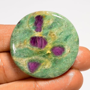 Peut inclure: Une pierre précieuse ronde et polie, de couleur verte avec des inclusions violettes vibrantes. La pierre présente une texture tachetée avec des zones jaunes et brunes. Le bijou est tenu dans une main, mettant en valeur sa beauté naturelle.