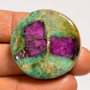 May include: A round gemstone with a vibrant mix of colors. The stone features patches of deep magenta within a sea of green and gold. The polished surface highlights the natural patterns and textures of the mineral, creating a unique and eye-catching piece.