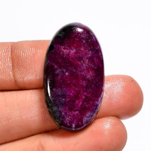 Könnte beinhalten: Ein ovaler Edelstein mit polierter Oberfläche. Der Stein hat eine tiefe magentafarbene Farbe mit helleren violetten und weißen Adern. Dunklere Bereiche in Schwarz und Grün sind oben und unten am Stein sichtbar.