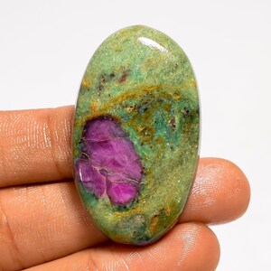 Puede incluir: Gema ovalada con una vibrante mezcla de colores. La piedra presenta un llamativo centro magenta rodeado por una matriz moteada verde y dorada. La superficie pulida refleja la luz, resaltando los patrones naturales.