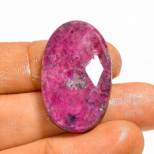 May include: A polished, oval gemstone in a vivid pink shade. The stone has a glossy surface with some lighter inclusions and a diamond-shaped highlight. The gemstone is held in a hand, showing its size and colour. The stone is likely for jewellery.