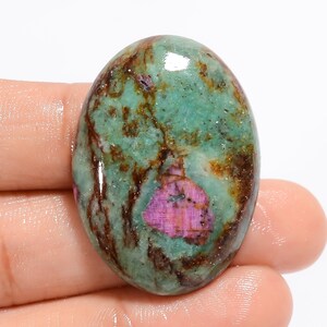 Könnte beinhalten: Oval geformter Edelstein mit einer Mischung aus Farben. Der Stein hat eine Basis aus Türkis und Grün mit braunen Adern und einem markanten rosa-roten Einschluss. Die polierte Oberfläche reflektiert das Licht und hebt die natürlichen Muster hervor.