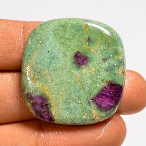 May include: A polished, square-shaped gemstone with a light green base color. The stone features several deep purple inclusions and a glossy finish. The gemstone is held in a hand.