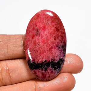May include: An oval gemstone, predominantly pink with a darker base. A striking black band cuts across the lower section. The polished surface of the stone reflects light, showcasing its natural texture and variations in colour.