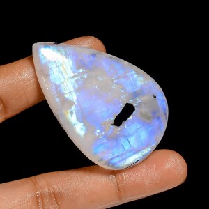 May include: A polished, pear-shaped moonstone gemstone, displaying a milky white base with iridescent blue and yellow tones. A black inclusion is visible within the stone. The gemstone is held between fingers against a black backdrop.