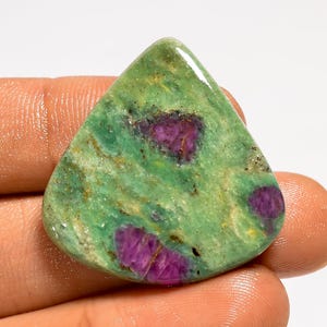 May include: A polished, triangular gemstone with a vibrant green base color. The stone features irregular patches of deep magenta and subtle brown inclusions, creating a unique pattern. The gem is held in a hand.