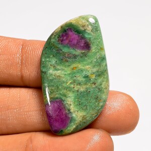 Puede incluir: Una piedra preciosa pulida con una base verde y vibrantes inclusiones magenta. La piedra, de forma suave y curvada, se sostiene en una mano. La superficie de la piedra preciosa tiene un aspecto natural y texturizado, con variaciones de color y patrón.