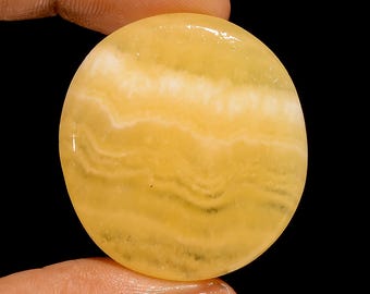 Puede incluir: Una gema redonda y pulida con un patrón de bandas amarillas y blancas. La piedra se sostiene entre los dedos sobre un fondo negro, mostrando sus variaciones de color naturales y su superficie lisa. El diseño de la gema recuerda a las formaciones naturales.
