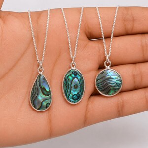 May include: Three silver necklaces featuring iridescent abalone shell pendants in teardrop, oval, and circular shapes. Each pendant is encased in a smooth silver bezel setting and hangs from a delicate silver chain, showcasing deep green and blue hues.