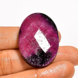 May include: An oval-cut gemstone, predominantly magenta with black inclusions. The polished surface features a large, reflective facet. The gem is held in a hand, highlighting its colour and shape. A striking piece of jewellery.