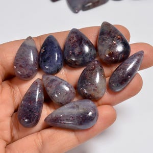 Puede incluir: Una colección de nueve piedras preciosas pulidas en forma de lágrima, con un color base azul y morado intenso y motas de inclusiones de color rojo oscuro y metálico en las superficies lisas y reflectantes de cada pieza de piedra.