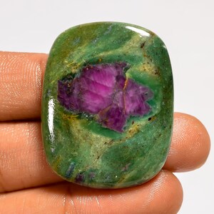 Può includere: Una gemma lucidata con una base verde vibrante e un'inclusione centrale di colore magenta intenso. La pietra ha una forma rettangolare arrotondata e una finitura lucida, che mostra motivi naturali e variazioni di colore.