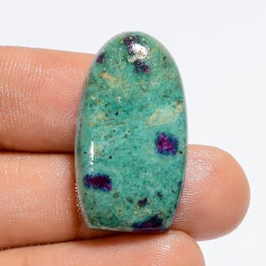 May include: Oval-shaped green gemstone cabochon with red and purple inclusions. The stone has a mottled, marbled appearance.