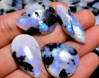 Puede incluir: Cuatro cabujones de piedra lunar iridiscente con inclusiones negras. Las piedras tienen forma de pera y tienen un brillo blanco, azul y morado.