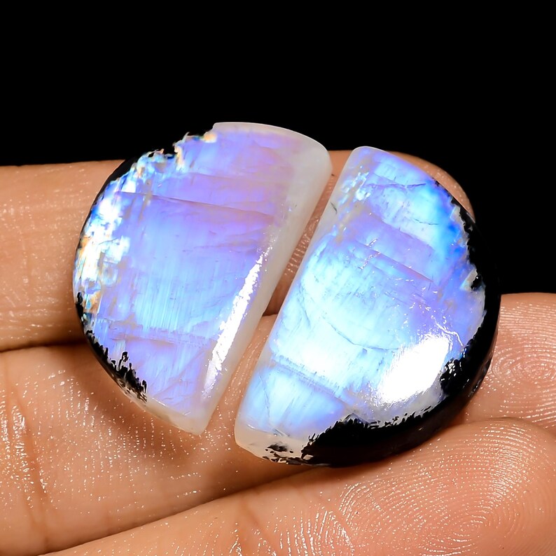 May include: Two polished moonstone cabochons, each a semi-circle shape. The stones display a milky white base with iridescent blue and purple flashes. The edges are outlined with a dark, almost black, border. The stones are set against a dark background.