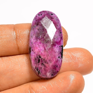May include: An oval gemstone with a faceted top, held in a hand. The stone is a vivid pink with black and grey inclusions. The polished surface highlights the natural variations. This gemstone, likely for jewellery, measures approximately 3 cm in length.