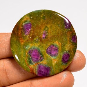 Peut inclure: Une pierre précieuse ronde et polie, présentant un mélange de couleurs vives. La base est vert olive et brun doré, parsemée d'inclusions magenta foncé. La surface polie reflète la lumière, mettant en valeur la beauté naturelle de la pierre.
