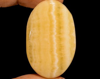 Puede incluir: Gema ovalada de color amarillo cálido con sutiles rayas blancas y amarillo claro. La piedra pulida se sostiene entre los dedos sobre un fondo negro, mostrando sus patrones naturales y variaciones de color. Una pieza única para la fabricación de joyas.