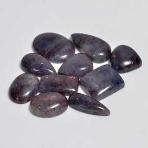 Puede incluir: Una colección de nueve piedras preciosas pulidas en corte cabujón con formas variadas, incluyendo óvalos, peras y rectángulos. Las piedras presentan un color base púrpura y gris profundo y translúcido con inclusiones rojizas y marrones brillantes.