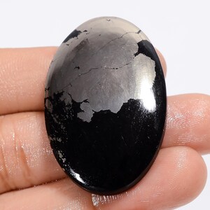 Pode incluir: Uma pedra preciosa oval cabochão de cor preta e prateada com uma superfície polida. A pedra tem um padrão único de inclusões de pirita prateada.