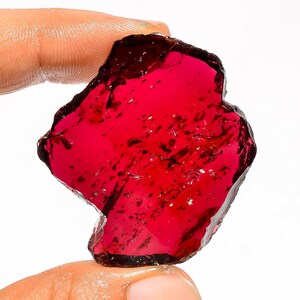 May include: A vibrant, raw garnet gemstone with a deep red hue. The rough-cut stone has a natural, irregular shape and a slightly translucent appearance. Darker inclusions are visible within the stone, adding to its unique character. The gemstone is held between two fingers.
