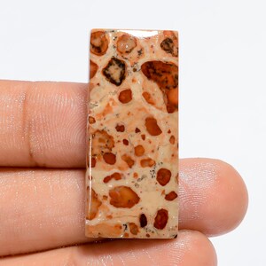 May include: A rectangular, light brown and orange gemstone cabochon with a mottled pattern. The stone has a polished surface and is set against a white background.