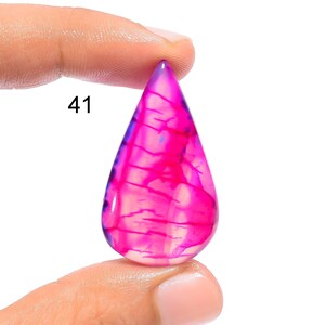 Puede incluir: Una piedra preciosa de ágata de forma de lágrima, rosa y púrpura, con un fondo blanco y patrones intrincados. La piedra preciosa está siendo sostenida por un dedo con el número 41 visible.