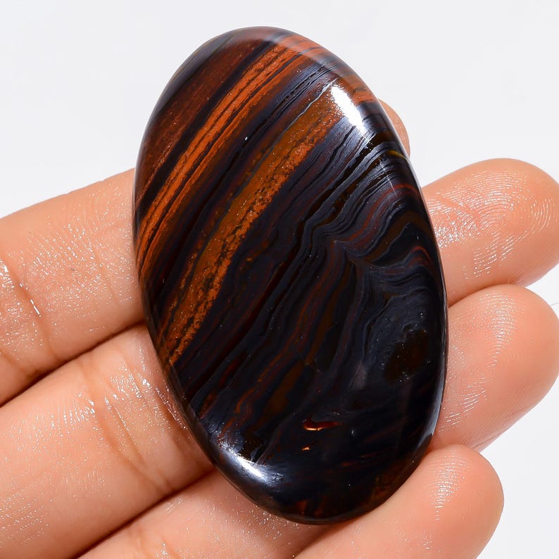 May include: Oval-shaped polished gemstone with a striking pattern of dark brown, black, and reddish-orange bands. The stone is held in a hand, showcasing its smooth, glossy surface and natural color variations. A unique, eye-catching piece.