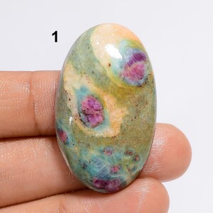 Könnte beinhalten: Ein ovaler Edelstein mit einer Mischung aus Farben. Der Stein hat eine Basis aus grünen und blauen Farbtönen mit Flecken in Rosa und Creme. Der Edelstein hat eine polierte Oberfläche und wird in einer Hand gehalten.