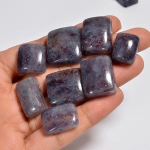 Puede incluir: Una colección de piedras pulidas y rodadas en tonos púrpura oscuro y gris, con motas rojas oscuras y metálicas dispersas por sus superficies lisas y redondeadas. Estas pequeñas piezas rectangulares irregulares tienen un acabado brillante.