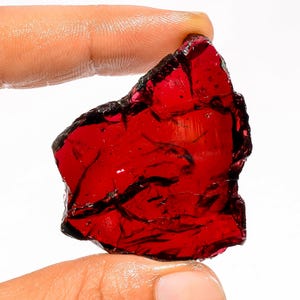 May include: A raw, translucent gemstone with a deep red hue. The stone has a rough, uneven surface and a dark, almost black edge. The gemstone is held between two fingers against a white background.