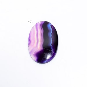 May include: A purple, white, and blue agate cabochon with a swirling pattern. The cabochon is oval shaped and has a smooth, polished surface.