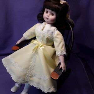 K&ouml;nnte beinhalten: Eine Porzellanpuppe mit braunen Haaren und einem gelben Kleid mit Spitzenbesatz sitzt in einem h&ouml;lzernen Schaukelstuhl. Die Puppe hat braune Augen und einen wei&szlig;en Kragen.