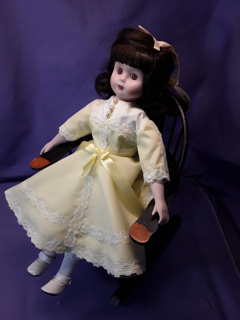 K&ouml;nnte beinhalten: Eine Porzellanpuppe mit braunen Haaren und einem gelben Kleid mit Spitzenbesatz sitzt in einem h&ouml;lzernen Schaukelstuhl. Die Puppe hat braune Augen und einen wei&szlig;en Kragen.