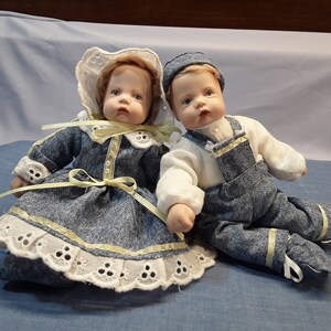K&ouml;nnte beinhalten: Zwei Porzellanpuppen in Denim-Outfits. Die M&auml;dchenpuppe tr&auml;gt eine wei&szlig;e Haube und ein gelbes Band. Die Jungenpuppe tr&auml;gt eine blaue M&uuml;tze und ein gelbes Band an ihrer Latzhose.
