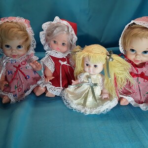 K&ouml;nnte beinhalten: Vier Vintage-Babypuppen mit blonden und wei&szlig;en Haaren, die rote und wei&szlig;e Gingham-Kleider mit Spitzenbesatz und H&auml;ubchen tragen.