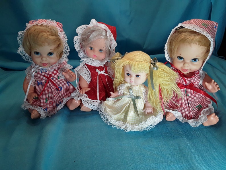 K&ouml;nnte beinhalten: Vier Vintage-Babypuppen mit blonden und wei&szlig;en Haaren, die rote und wei&szlig;e Gingham-Kleider mit Spitzenbesatz und H&auml;ubchen tragen.