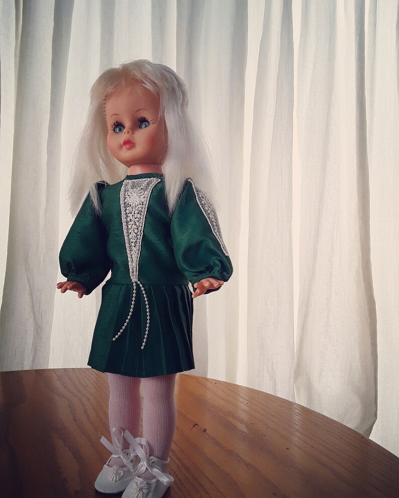 K&ouml;nnte beinhalten: Eine Vintage-Puppe mit wei&szlig;en Haaren und blauen Augen, die ein gr&uuml;nes Kleid mit wei&szlig;en Spitzenbesatz und wei&szlig;en Schuhen tr&auml;gt.