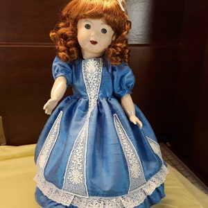 K&ouml;nnte beinhalten: Eine Porzellanpuppe mit roten Locken, die ein blaues Kleid mit wei&szlig;en Spitzenbesatz tr&auml;gt.