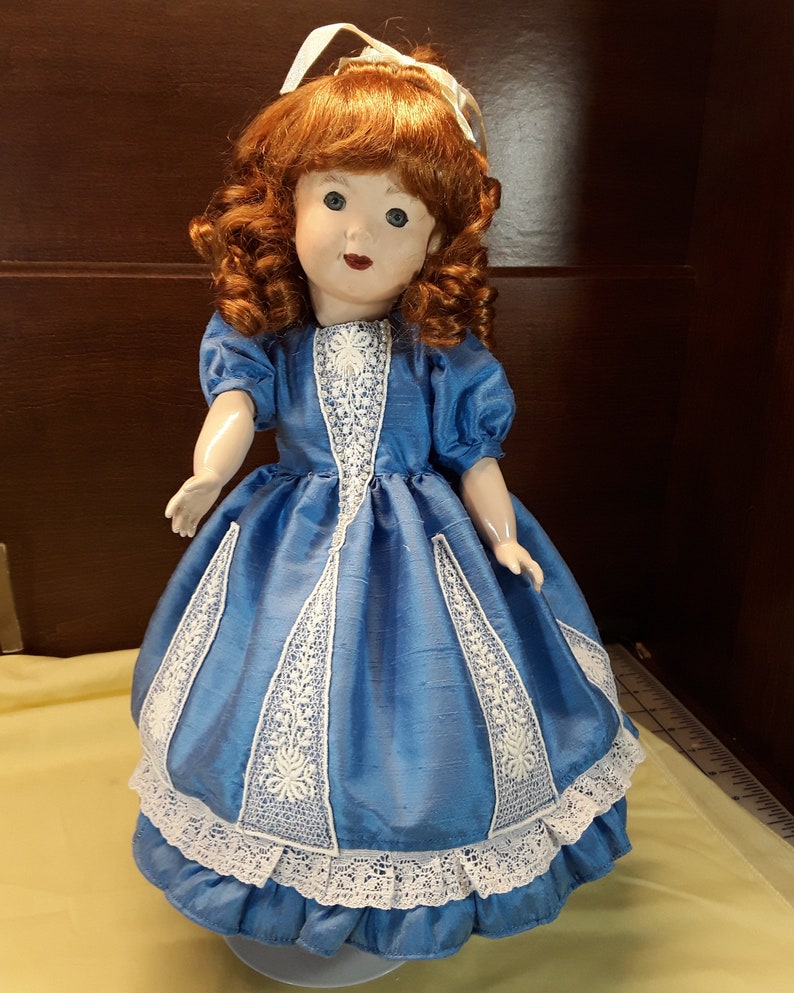 K&ouml;nnte beinhalten: Eine Porzellanpuppe mit roten Locken, die ein blaues Kleid mit wei&szlig;en Spitzenbesatz tr&auml;gt.