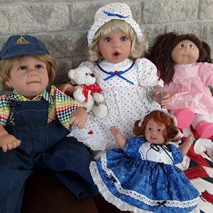 K&ouml;nnte beinhalten: Vier Puppen, zwei mit blonden Haaren, eine mit braunen Haaren und eine mit roten Haaren. Die Puppen tragen verschiedene Outfits, darunter ein blaues und wei&szlig;es gepunktetes Kleid, ein rosa Kleid, ein blaues und wei&szlig;es Kleid und eine Latzhose mit einem Regenbogenhemd. Eine Puppe h&auml;lt einen Teddyb&auml;r.