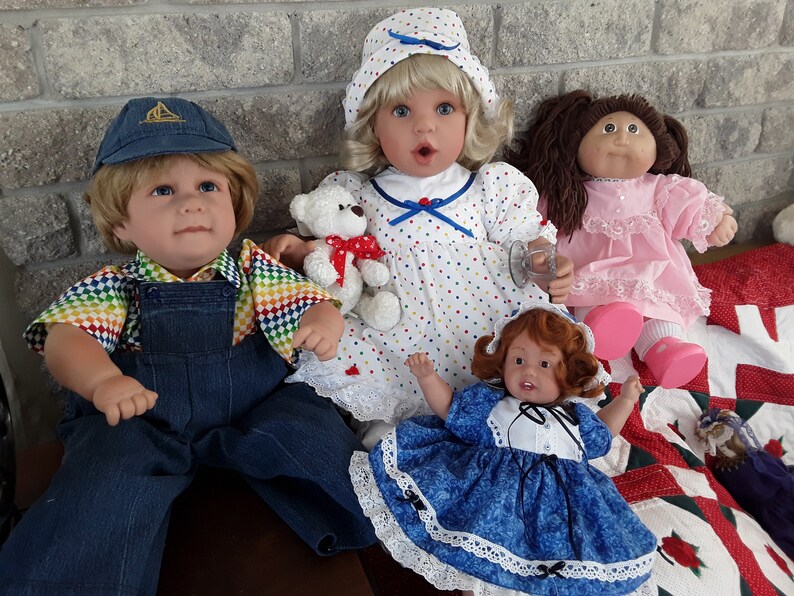 K&ouml;nnte beinhalten: Vier Puppen, zwei mit blonden Haaren, eine mit braunen Haaren und eine mit roten Haaren. Die Puppen tragen verschiedene Outfits, darunter ein blaues und wei&szlig;es gepunktetes Kleid, ein rosa Kleid, ein blaues und wei&szlig;es Kleid und eine Latzhose mit einem Regenbogenhemd. Eine Puppe h&auml;lt einen Teddyb&auml;r.