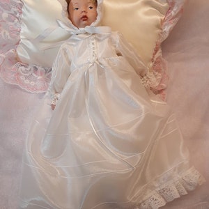 K&ouml;nnte beinhalten: Eine wei&szlig;e Porzellan-Babypuppe, die ein wei&szlig;es Satin-Taufkleid mit Spitzenbesatz tr&auml;gt. Die Puppe liegt auf einem wei&szlig;en Satin-Kissen mit rosa Spitzenbesatz.