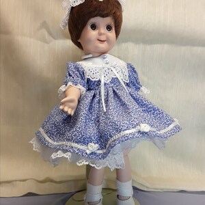 K&ouml;nnte beinhalten: Eine Porzellanpuppe, die ein blau-wei&szlig;es Blumenkleid mit wei&szlig;en Spitzenbesatz tr&auml;gt. Die Puppe hat braunes Haar und blaue Augen und tr&auml;gt wei&szlig;e Schuhe.