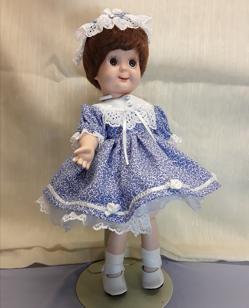 K&ouml;nnte beinhalten: Eine Porzellanpuppe, die ein blau-wei&szlig;es Blumenkleid mit wei&szlig;en Spitzenbesatz tr&auml;gt. Die Puppe hat braunes Haar und blaue Augen und tr&auml;gt wei&szlig;e Schuhe.