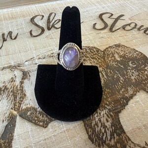 May include: A silver ring featuring an oval-shaped, polished purple and white stone set in a metal bezel. The band has decorative etched lines on the sides near the stone setting. The ring is displayed on a black velvet cone-shaped stand.