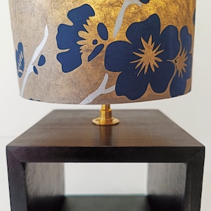 Könnte beinhalten: Eine goldene und schwarze Tischlampe mit einem floralen Lampenschirm. Der Lampenschirm zeigt blaue Blumen mit goldenen Akzenten auf goldenem Hintergrund.